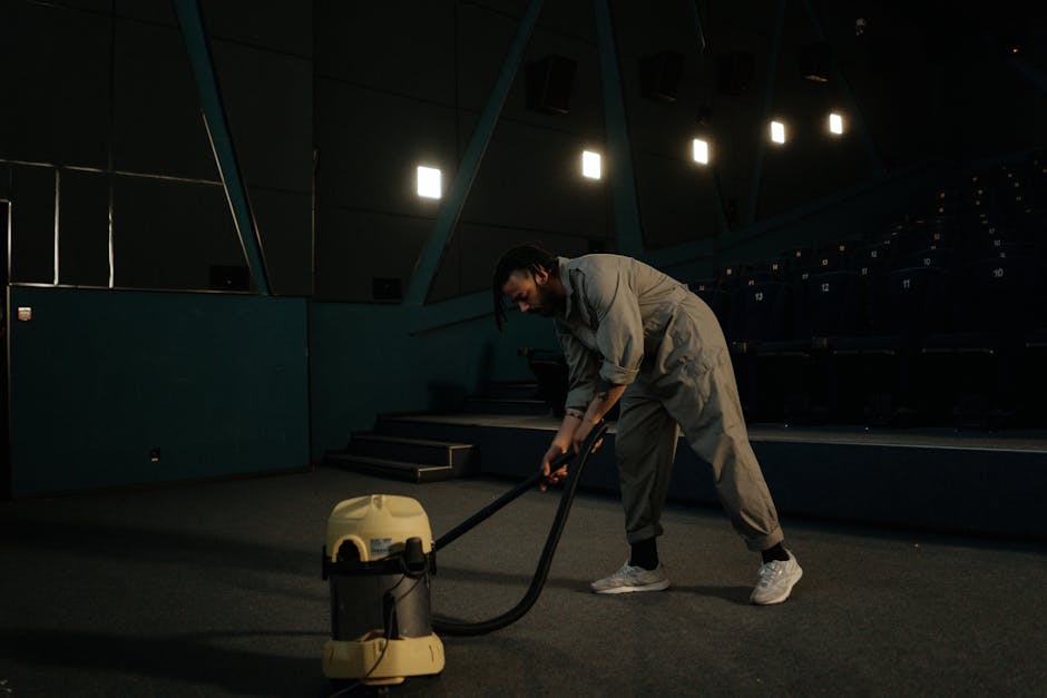 A woman with curly hair wearing a beige jacket, blue jeans, and white shoes is conducting surface cleaning using a yellow vacuum cleaner with a black hose and a cleaning wand. She is standing on a pat