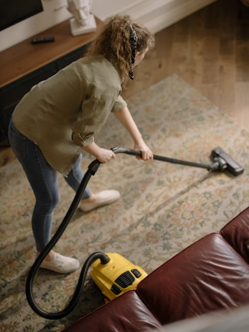A woman with curly hair, dressed in a light-colored shirt, jeans, and white sneakers, is engaged in surface cleaning with a vacuum cleaner in a living room. She is holding the vacuum hose with both ha